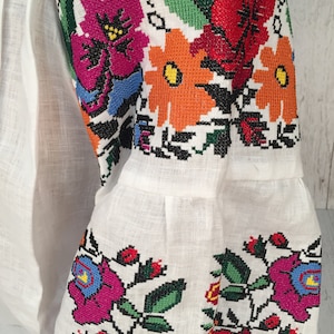 May include: White linen blouse with colorful embroidered floral patterns on the sleeves. The embroidery features red, orange, yellow, green, blue, and pink flowers.