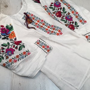 May include: White linen blouse with colorful embroidered floral patterns on the sleeves and neckline.