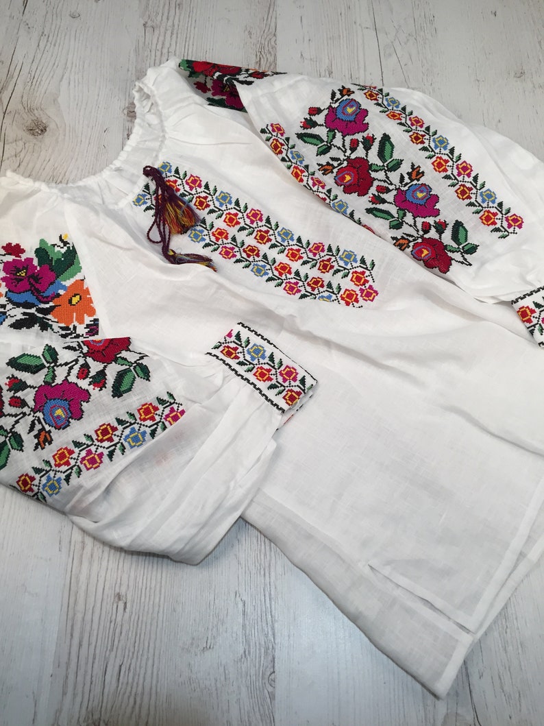 May include: White linen blouse with colorful embroidered floral patterns on the sleeves and neckline.