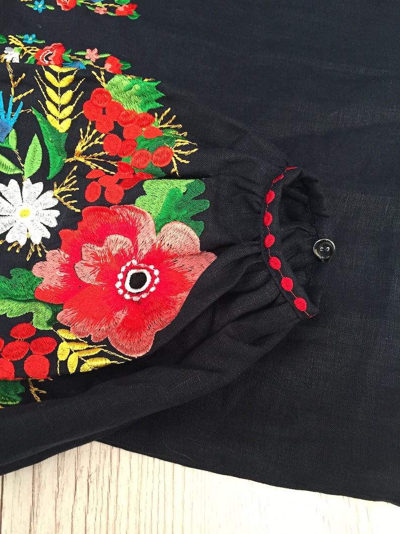 May include: A black linen blouse with red and green embroidered flowers. The embroidery is detailed and intricate, featuring a variety of floral motifs.