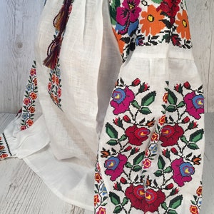 May include: White linen blouse with colorful embroidered floral patterns on the sleeves and neckline. The embroidery features red, pink, blue, green, orange, and yellow flowers.
