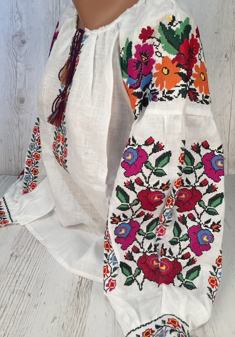 May include: White linen blouse with colorful embroidered floral patterns on the sleeves and neckline. The embroidery features red, pink, blue, green, orange, and yellow flowers.