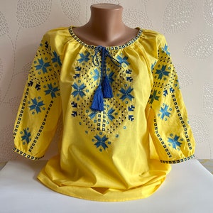 May include: A yellow embroidered blouse with blue and black floral patterns. The blouse has a tie at the neckline with blue tassels.