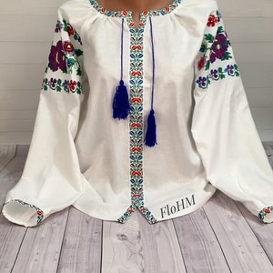 May include: White linen blouse with embroidered floral patterns in red, blue, and green. The blouse has a V-neckline and long sleeves with blue tassels.