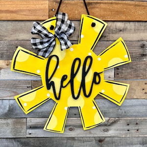 K&ouml;nnte beinhalten: Ein gelber, sonnenf&ouml;rmiger T&uuml;rkranz mit wei&szlig;en Punkten und dem Wort "hello" in schwarzer Schrift. Eine schwarz-wei&szlig; karierte Schleife ist oben angebracht.