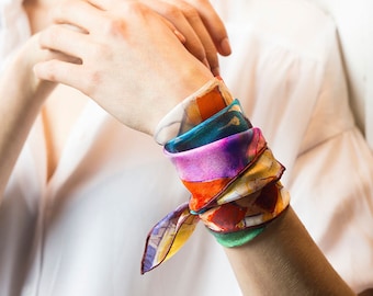 May include: A colorful silk scarf tied around a wrist as a bracelet. The scarf has a watercolor-like design with shades of yellow, orange, red, green, blue, and purple.