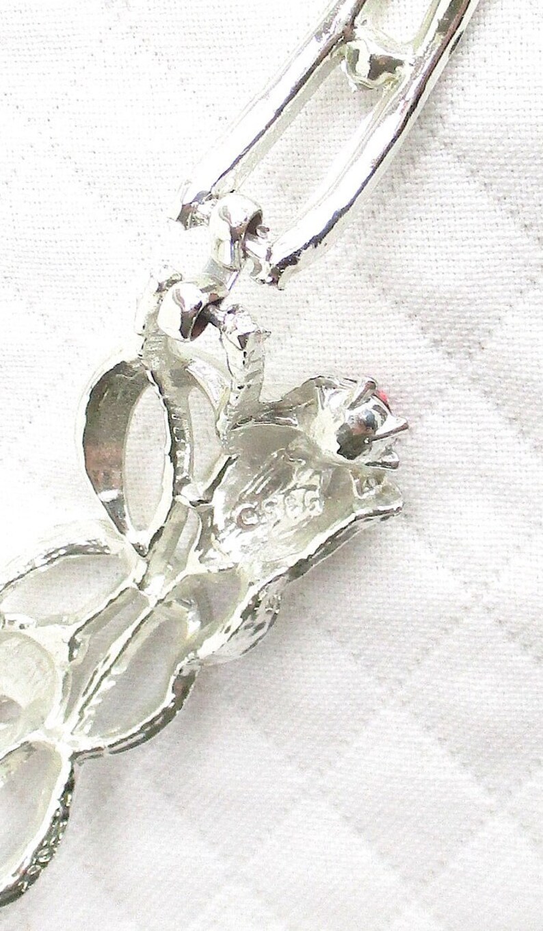 Puede incluir: Broche plateado con dise&ntilde;o floral y una peque&ntilde;a gema roja. El broche tiene una superficie texturizada y un cierre de alfiler. El dise&ntilde;o incluye un patr&oacute;n de hojas y un lazo.