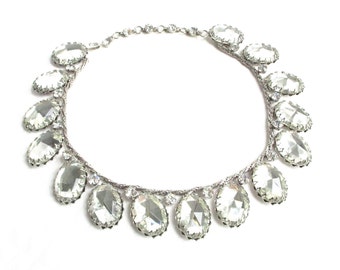 May include: A silver necklace with a row of large, clear, faceted crystal stones. The stones are oval shaped and have a silver setting.