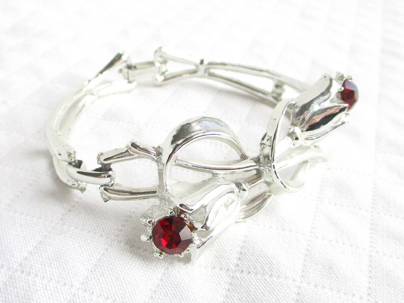 Puede incluir: Pulsera de metal plateado con dise&ntilde;o floral. Presenta tres elementos en forma de tulip&aacute;n, cada uno con una gema roja vibrante. La pulsera articulada parece estar hecha de metal ligero.
