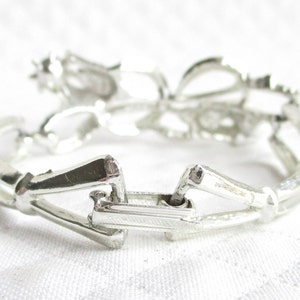 Puede incluir: Un brazalete plateado con un dise&ntilde;o floral. El brazalete es de metal y tiene un cierre de broche. Los elementos florales son visibles en la parte superior. El brazalete est&aacute; sobre una superficie blanca.