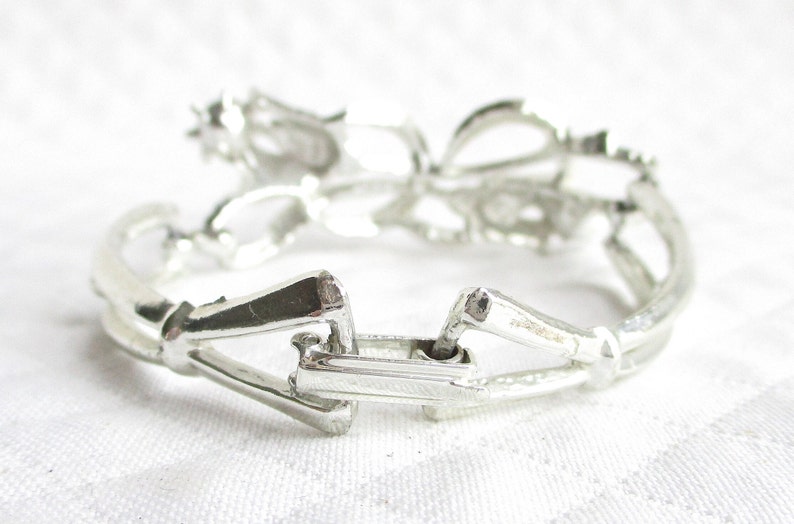 Puede incluir: Un brazalete plateado con un dise&ntilde;o floral. El brazalete es de metal y tiene un cierre de broche. Los elementos florales son visibles en la parte superior. El brazalete est&aacute; sobre una superficie blanca.