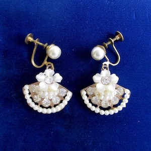 Puede incluir: Un par de pendientes vintage de clip. Cada pendiente presenta un perno de perla, una flor blanca con un pedrería central y un diseño en forma de abanico con perlas de imitación y pedrería transparente. Los pendientes tienen un acabado metálico dorado.