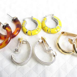 May include: A set of six earrings, including two pairs of yellow hoop earrings, one pair of brown hoop earrings, and two pairs of silver clip-on earrings.