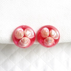 May include: Pair of round, red resin earrings. Each earring features three small, pearlescent seashells encased in clear resin. The earrings are set against a white, textured background.