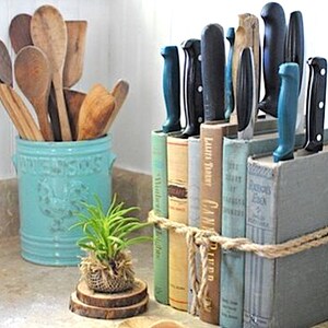 May include: A set of kitchen knives are stored in a vertical book holder made of old hardback books. The books are tied together with twine. The knives are all different sizes and have blue handles. The book holder is sitting on a counter top next to a blue ceramic utensil holder filled with wooden spoons.