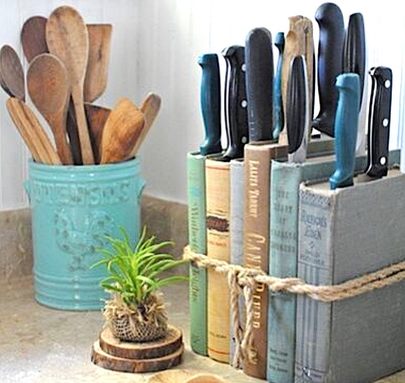 May include: A set of kitchen knives are stored in a vertical book holder made of old hardback books. The books are tied together with twine. The knives are all different sizes and have blue handles. The book holder is sitting on a counter top next to a blue ceramic utensil holder filled with wooden spoons.