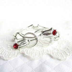 Puede incluir: Pulsera plateada con dise&ntilde;o floral, con tres elementos en forma de tulip&aacute;n. Cada tulip&aacute;n tiene una gema roja brillante en el centro. La pulsera est&aacute; sobre un fondo blanco texturizado.
