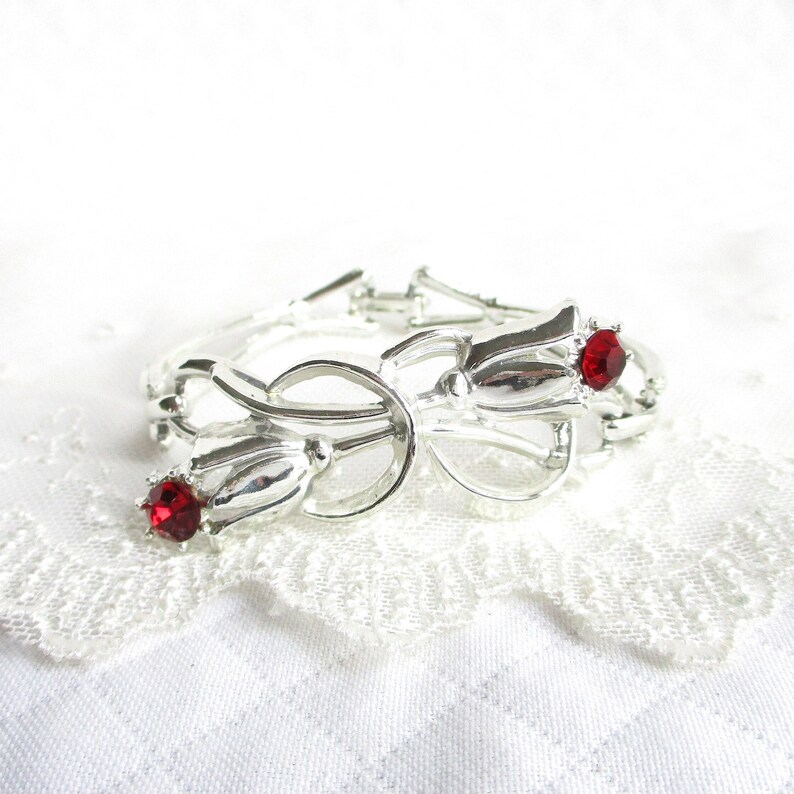 Puede incluir: Pulsera plateada con dise&ntilde;o floral, con tres elementos en forma de tulip&aacute;n. Cada tulip&aacute;n tiene una gema roja brillante en el centro. La pulsera est&aacute; sobre un fondo blanco texturizado.