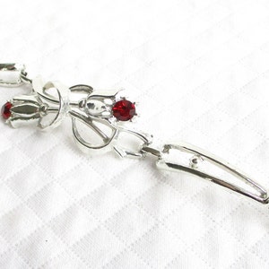 Puede incluir: Broche plateado con dise&ntilde;o floral. El broche presenta tres gemas rojas. El broche est&aacute; hecho de un material met&aacute;lico brillante y se coloca sobre un fondo blanco.
