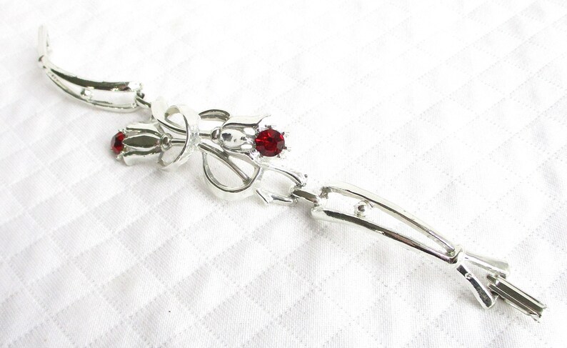 Puede incluir: Broche plateado con dise&ntilde;o floral. El broche presenta tres gemas rojas. El broche est&aacute; hecho de un material met&aacute;lico brillante y se coloca sobre un fondo blanco.