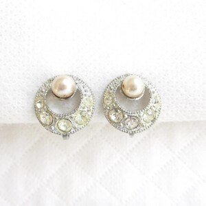 May include: A pair of vintage clip-on earrings. Each earring features a silver-toned, circular design with a faux pearl center and surrounding clear rhinestones. The earrings are set against a white background.
