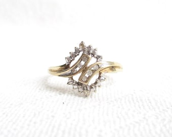 May include: A gold-toned ring with a cluster of small, clear gemstones. The ring features a unique design with a central, curved section and a surrounding halo of stones. The band is a simple, polished gold. The ring is set against a white background.