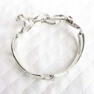 Puede incluir: Pulsera articulada plateada con dise&ntilde;o floral. La pulsera presenta tres adornos en forma de flor, cada uno con una peque&ntilde;a gema roja en el centro. La pulsera est&aacute; hecha de un material met&aacute;lico brillante y tiene un cierre de broche.