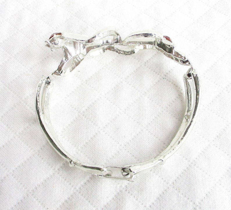 Puede incluir: Pulsera articulada plateada con dise&ntilde;o floral. La pulsera presenta tres adornos en forma de flor, cada uno con una peque&ntilde;a gema roja en el centro. La pulsera est&aacute; hecha de un material met&aacute;lico brillante y tiene un cierre de broche.