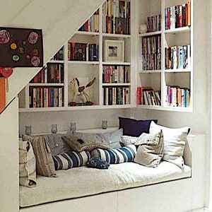 May include: A white built-in bookshelf with a white daybed underneath. The bookshelf is filled with books and has a white rocking horse on the bottom shelf. The daybed has a white mattress and is covered with pillows in various shades of blue and beige.