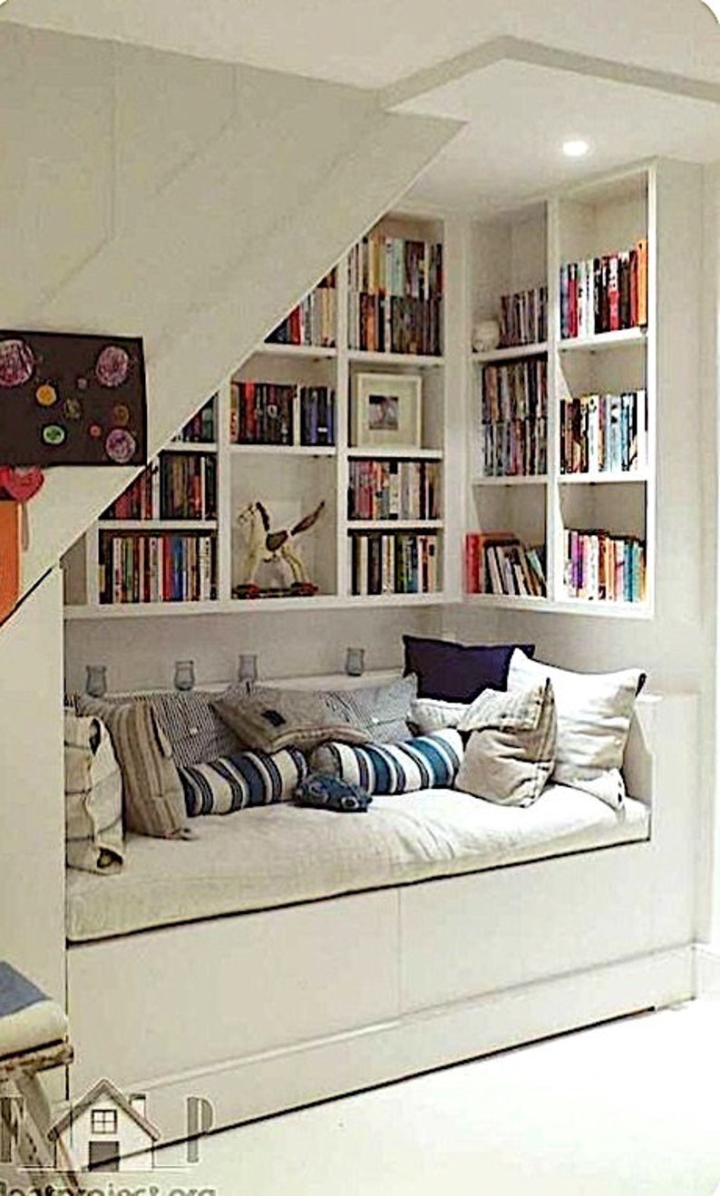 May include: A white built-in bookshelf with a white daybed underneath. The bookshelf is filled with books and has a white rocking horse on the bottom shelf. The daybed has a white mattress and is covered with pillows in various shades of blue and beige.