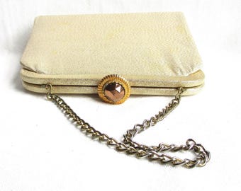 May include: A vintage, rectangular clutch purse in a light gold color. The purse features a textured surface, a decorative gold-toned clasp with a brown gem, and a gold chain strap. The purse is closed.