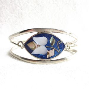 Peut inclure: Un bracelet manchette en argent avec une incrustation ovale centrale en résine bleu profond, agrémentée de fragments de coquillages blancs irisés et nacrés. La bande métallique est lisse avec des détails en fil torsadé de chaque côté.