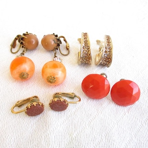 May include: Assortment of vintage clip-on earrings with various colored stones. The earrings feature a variety of styles, including round, oval, and hoop shapes. The stones are a mix of orange, red, and brown.