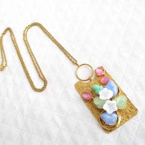 Op de afbeelding: Goudkleurige ketting met een rechthoekige hanger met kleurrijke bloemaccenten. De hanger heeft een textuur en is versierd met roze, blauwe, groene en witte bloemvormige stenen. De ketting is lang en delicaat.