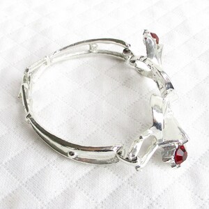 Puede incluir: Pulsera plateada con dise&ntilde;o floral y detalles de piedras preciosas rojas. La pulsera est&aacute; hecha de segmentos unidos y presenta un elemento decorativo con una piedra preciosa roja. La pulsera est&aacute; sobre una superficie blanca.