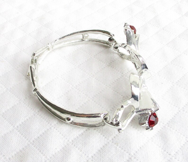 Puede incluir: Pulsera plateada con dise&ntilde;o floral y detalles de piedras preciosas rojas. La pulsera est&aacute; hecha de segmentos unidos y presenta un elemento decorativo con una piedra preciosa roja. La pulsera est&aacute; sobre una superficie blanca.