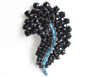 May include: A vintage brooch in a feather design. The brooch is made up of many black, faceted crystals, with a central stem of small, light blue beads. The brooch is set against a white background.