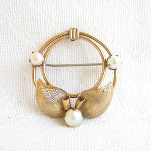 Puede incluir: Broche dorado vintage con diseño circular, con tres perlas de imitación y dos acentos de hojas estilizadas. El broche tiene un cierre de alfiler en la parte posterior y está sobre un fondo blanco.