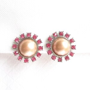 May include: A pair of vintage-style earrings. Each earring features a large, round, peach-colored center stone surrounded by a ring of small, pink, sparkling stones. The earrings have a silver-colored metal setting.