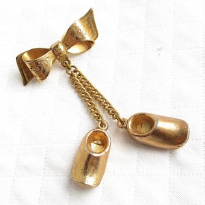 Könnte beinhalten: Goldfarbene Brosche mit Schleife und zwei kleinen Holzschuhen, die an Ketten hängen. Die Schleife hat geätzte Details, und die Holzschuhe sind hohl. Ein Schmuckstück im Vintage-Stil.