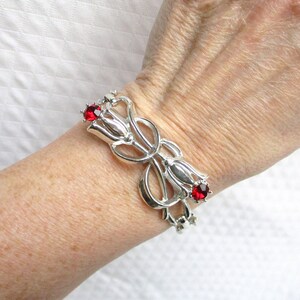 May include: A silver bracelet with a floral design, featuring three tulip-shaped elements. Each tulip has a red gemstone at its centre. The bracelet is worn on a wrist, highlighting its intricate details.