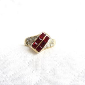 May include: A gold-toned ring with a rectangular red stone design. The ring features a central rectangular setting with two parallel red stones, flanked by smaller, clear stones. The band has a textured appearance.