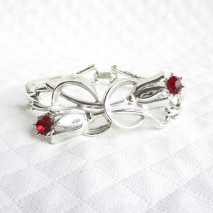 Puede incluir: Pulsera plateada con dise&ntilde;o floral y dos detalles de gemas rojas. La pulsera tiene un centro en forma de lazo y un cierre de broche. La pulsera se muestra sobre una superficie blanca texturizada.