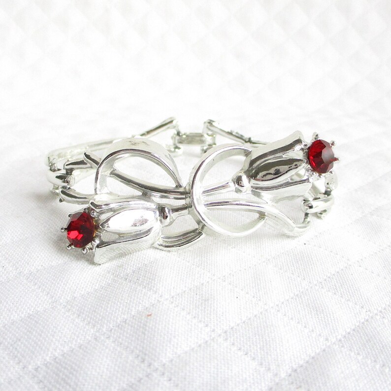 Puede incluir: Pulsera plateada con dise&ntilde;o floral y dos detalles de gemas rojas. La pulsera tiene un centro en forma de lazo y un cierre de broche. La pulsera se muestra sobre una superficie blanca texturizada.