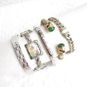 May include: A collection of vintage bracelets in silver and gold tones. Includes a silver bangle with a starburst pattern, a bracelet with a small framed picture, and a chain bracelet with green and blue accents.