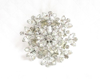 May include: A large, silver-toned brooch with a snowflake design. The brooch is made of clear rhinestones of various sizes and shapes.