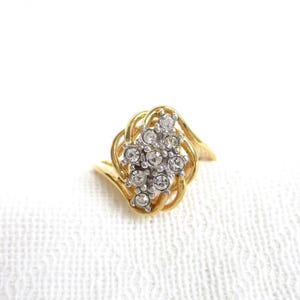 May include: A gold-toned ring featuring a cluster of clear, round gemstones. The ring has a decorative, swirling design around the gemstone cluster. The ring is set against a white background.