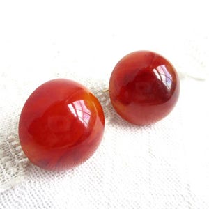 Könnte beinhalten: Ein Paar runde, rot-orangefarbene Ohrringe mit glänzendem Finish. Die Ohrringe sind vor einem weißen, strukturierten Hintergrund platziert. Sie haben ein glattes, poliertes Aussehen und reflektieren das Licht.