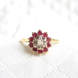 May include: A gold ring featuring a floral design. The ring's center stone is surrounded by a cluster of small, round, deep red gemstones. The center stone is a light-colored oval shape. The ring is set against a white background.