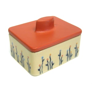Puede incluir: Una pequeña caja de cerámica con tapa. La caja está decorada con un patrón de flores azules y naranjas sobre un fondo crema. La tapa es de color naranja brillante y tiene una parte superior redondeada.
